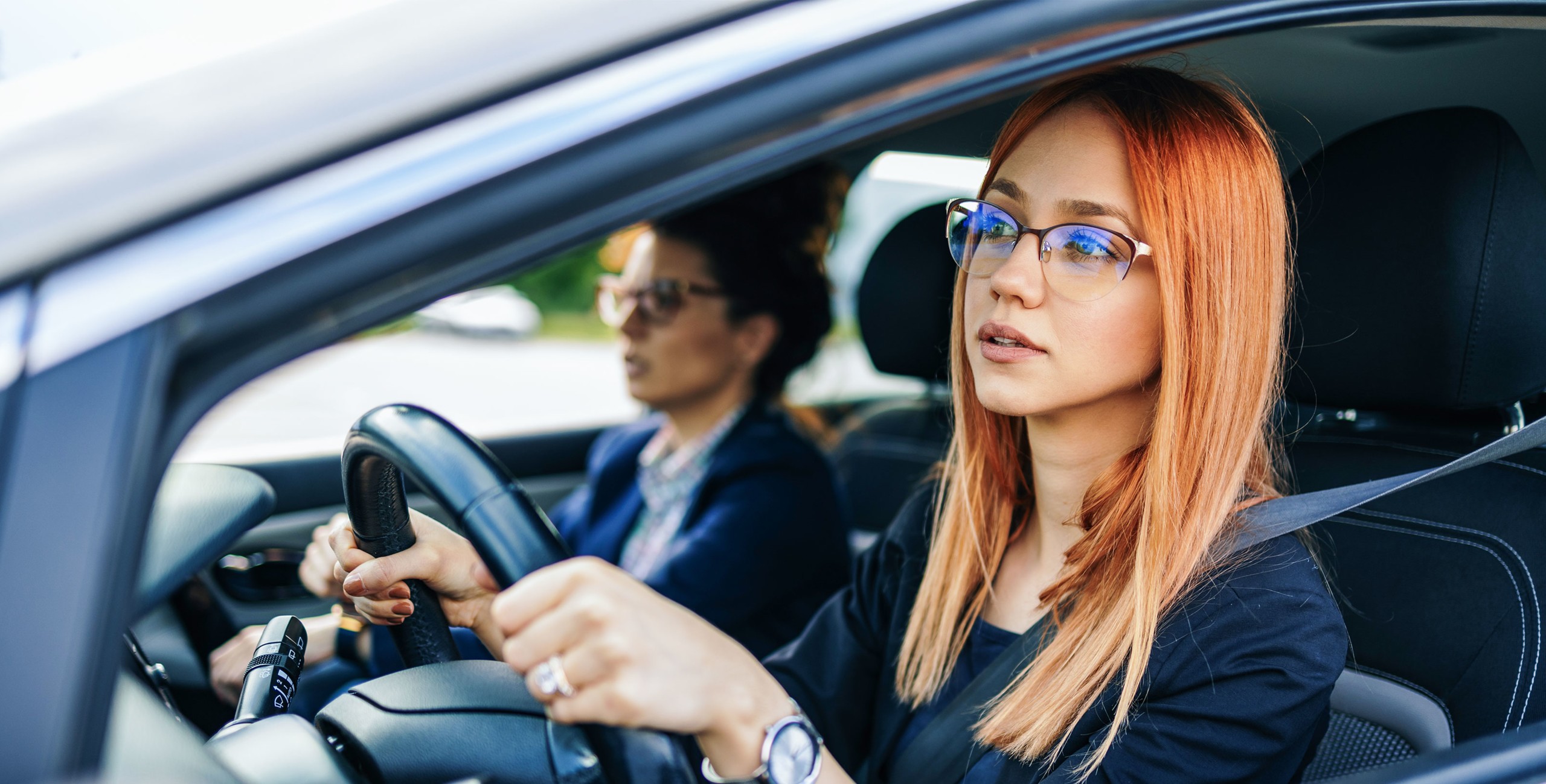Eine junge rothaarige Frau mit Brille sitzt am Steuer und schaut konzentriert auf die Strasse, während eine ältere Beifahrerin neben ihr sitzt.