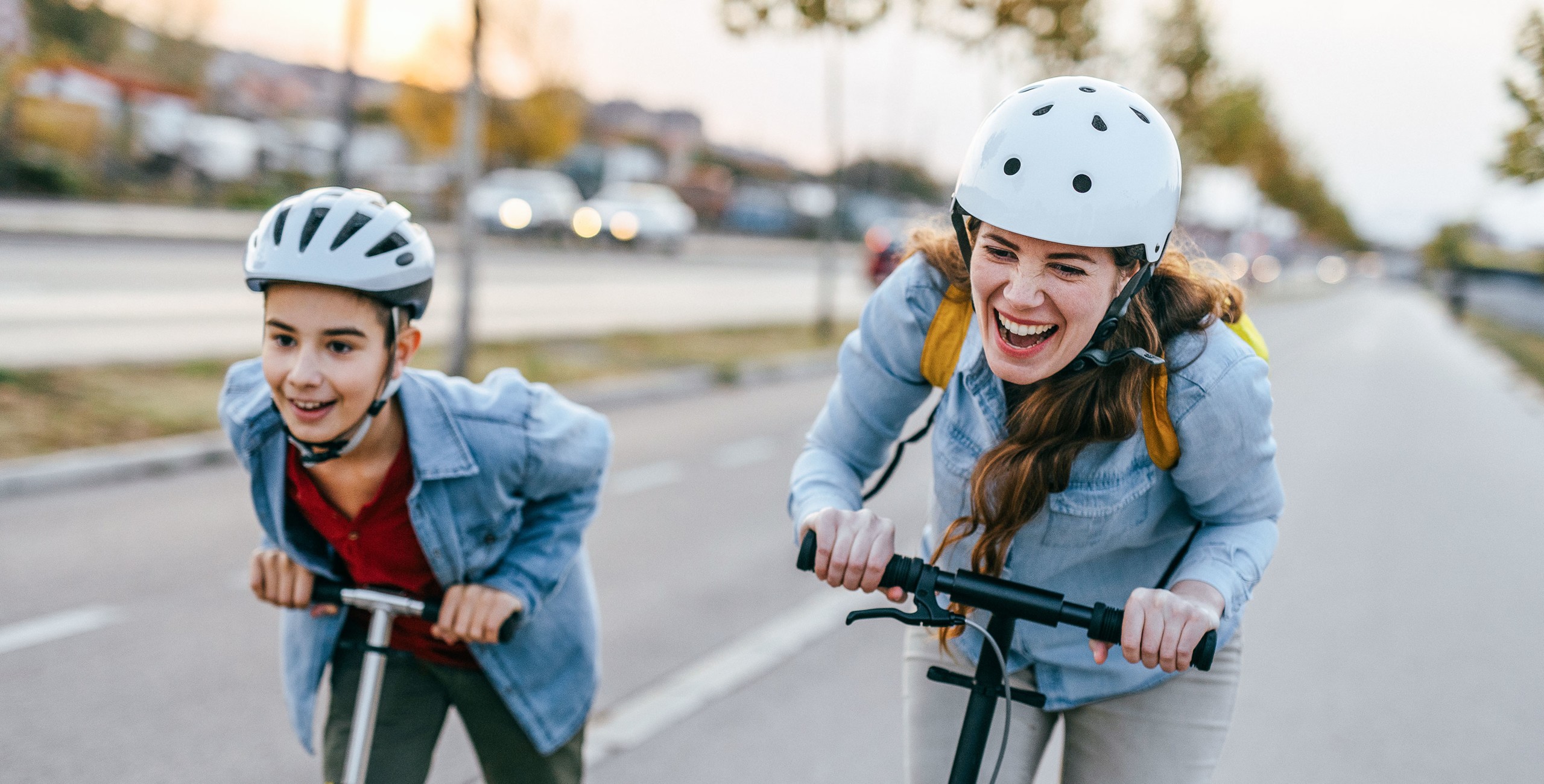 Deux jeunes portant un casque et vêtus d’une chemise en jean roulent en riant sur des scooters électriques sur une large route.