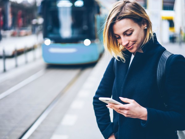 Une jeune femme en manteau noir au bord de la route regarde son téléphone en souriant, tandis qu’un tramway jaune s’approche à l’arrière-plan.