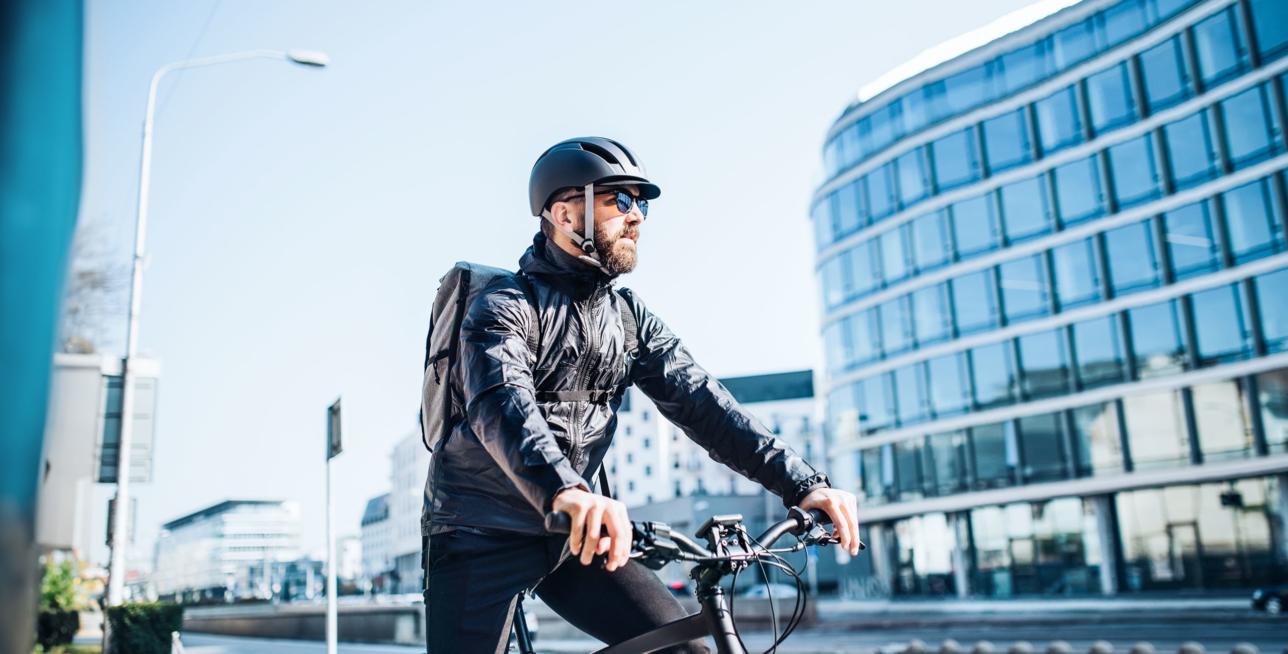 Ein Mann mit schwarzem Helm und Sonnenbrille steht mit seinem Fahrrad in einer städtischen Umgebung.