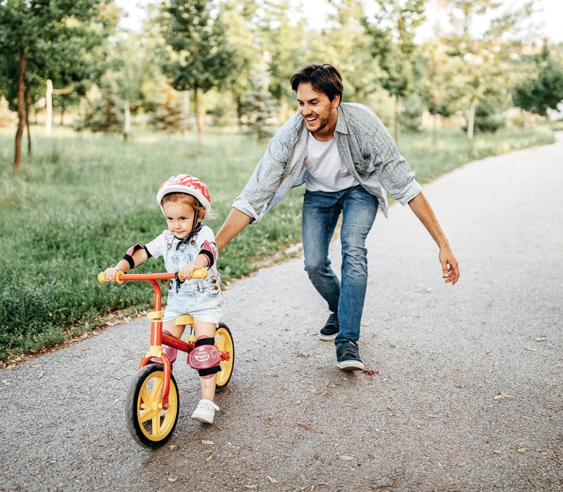 Un uomo sostiene una bimba con il casco che pedala sulla sua bicicletta lungo il viale di un parco.